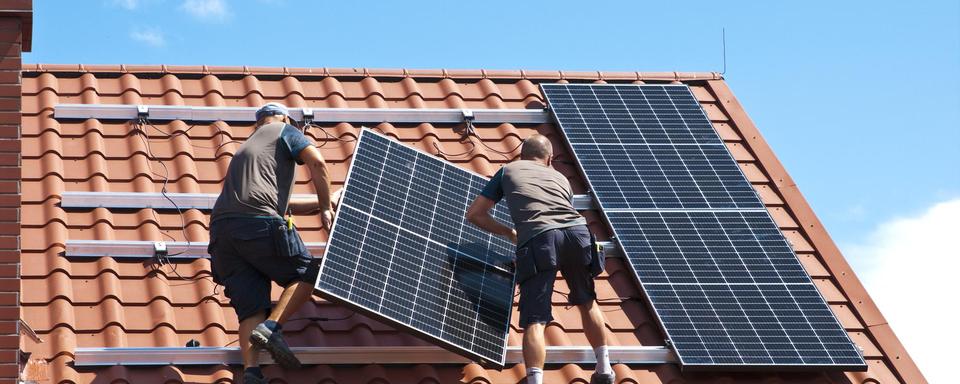 Deux ouvriers installent des panneaux solaires sur le toit d'une maison. [Depositphotos - AHatmaker]